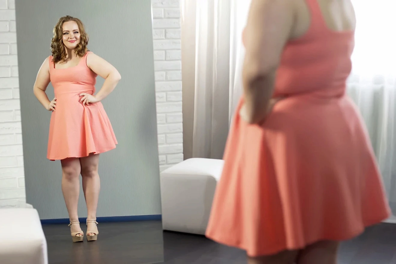 Cheerful plus size woman in dress looking in mirror